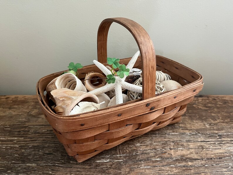 May include: A brown woven basket with a handle filled with white seashells, a white starfish, and green leaves.