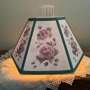 Könnte beinhalten: Ein sechseckiger Lampenschirm mit floralem Design. Der Schirm hat einen hellen Hintergrund mit gemalten lila Rosen und grünen Zierleisten. Ein klarer Glasschornstein befindet sich oben, und die Lampe wird von innen beleuchtet.