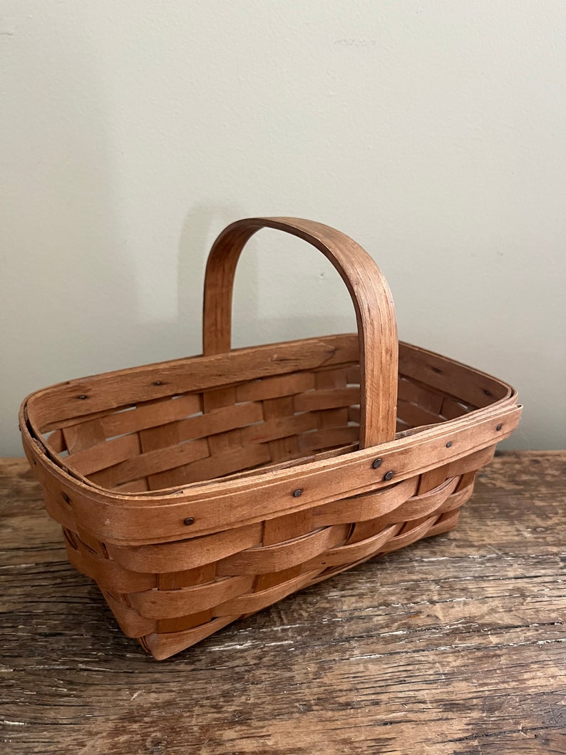 May include: A rectangular woven basket with a wooden handle. The basket is made of natural wood and has a light brown colour.
