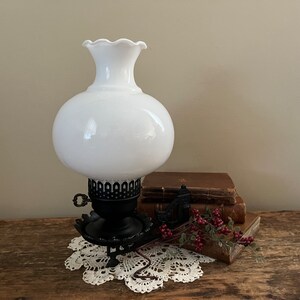 May include: A vintage white glass lamp with a round globe and a ruffled top. The lamp sits on a black metal base with a decorative design. A stack of old books and a sprig of red berries are next to the lamp. The lamp is on a white lace doily.