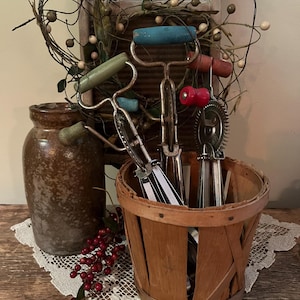May include: A set of vintage kitchen tools with wooden handles in a wooden basket. The tools include a whisk, a dough blender, and a pastry cutter. The tools are in a rustic setting with a brown crock and a wreath of greenery.