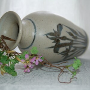 May include: A ceramic vase in a speckled beige finish, adorned with a blue band and stylized floral motifs. A sprig of artificial flowers with green leaves and pink blossoms rests near the vase.