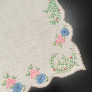 May include: Off-white linen placemat with a scalloped edge. The border features embroidered floral designs in pink and blue, with green vines and red berry accents. The placemat is a decorative item.