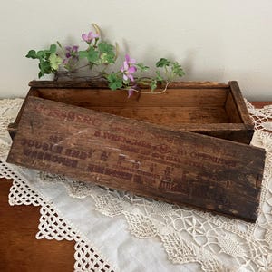 May include: An open, aged wooden box with a hinged lid, resting on a white lace doily. The lid features faded red text that reads "Double End Wrenches". A sprig of purple flowers and green leaves sits atop the box.