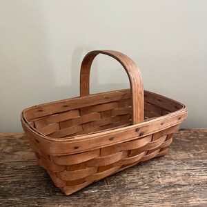 May include: A small, rectangular, wooden basket with a woven design and a single handle. The basket is made of natural wood and has a light brown colour.