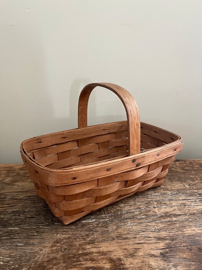 May include: A small, rectangular, wooden basket with a woven design and a single handle. The basket is made of natural wood and has a light brown colour.