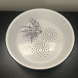 May include: White enamel colander with a black rim and a lavender sprig design. The colander has three sections of small drainage holes. This kitchenware is ideal for washing and draining fruits and vegetables.