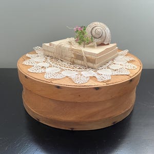 May include: A round, wooden box with a stack of books tied with twine, a seashell, and a doily. The box is light brown and has a lid. The books are beige, and the seashell is white. A small sprig of pink flowers sits atop the books.