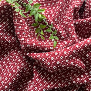 May include: A close-up of a textured, red fabric with a repeating white square pattern. The fabric is draped, creating folds and shadows. A green leafy sprig rests on top, adding a natural element to the composition.
