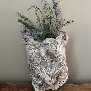 May include: White ceramic wall vase with a textured surface and a rope handle. The vase is filled with purple and green flowers.