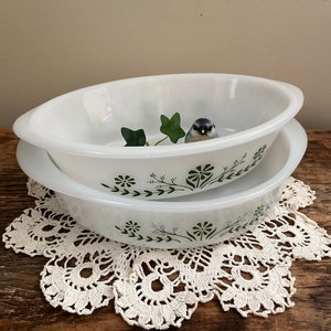 May include: Two white oval baking dishes with a green floral design and a small bird illustration. The dishes are stacked on top of each other and are sitting on a white crocheted doily.