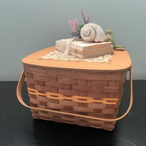 May include: A brown woven basket with a wooden lid and handle. The lid holds a stack of books, a seashell, a small bird figurine, and dried flowers. The basket has a decorative orange stripe.