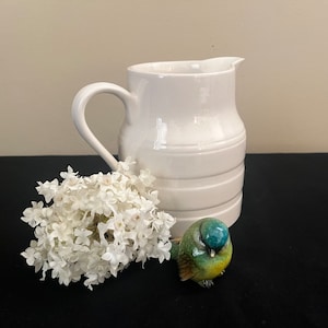 May include: A white ceramic pitcher with a wide mouth and a handle. The pitcher has a ribbed design and is sitting on a black surface. A small blue and green bird figurine is in front of the pitcher. A bouquet of white flowers is to the left of the pitcher.