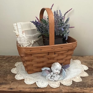 May include: A brown wicker basket filled with lavender flowers, a stack of white fabric, and a small porcelain angel figurine. The basket is sitting on a white crocheted doily on a wooden surface.
