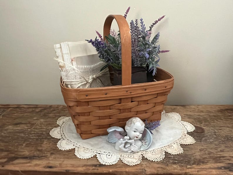 May include: A brown wicker basket filled with lavender flowers, a stack of white fabric, and a small porcelain angel figurine. The basket is sitting on a white crocheted doily on a wooden surface.