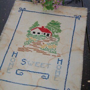 May include: White embroidered fabric with a blue border and a house with trees and flowers. The text "Home Sweet Home" is embroidered in blue.