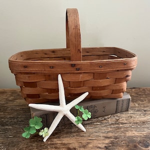 May include: A brown woven basket with a wooden handle sits on top of a book with a white starfish and green shamrocks on the front. The book is on a wooden surface.