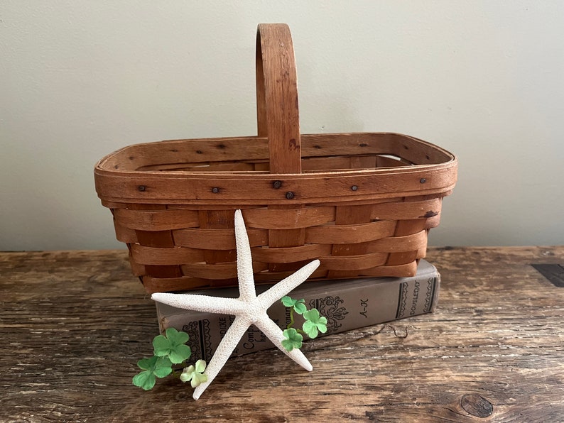 May include: A brown woven basket with a wooden handle sits on top of a book with a white starfish and green shamrocks on the front. The book is on a wooden surface.