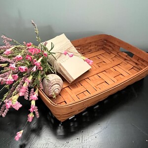 May include: A rectangular woven basket in a warm brown hue, with a bundle of blank tags tied with twine, a seashell, and a bouquet of pink and purple flowers. The basket has a handle on each side.