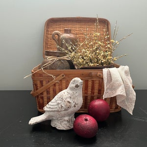 May include: A wicker picnic basket with a white bird figurine, a brown jug, a bouquet of white flowers, and two red apples. The basket is open and the items are arranged inside.