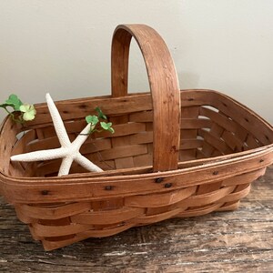 May include: A brown woven basket with a wooden handle. The basket is empty and has a white starfish and green leaves inside.