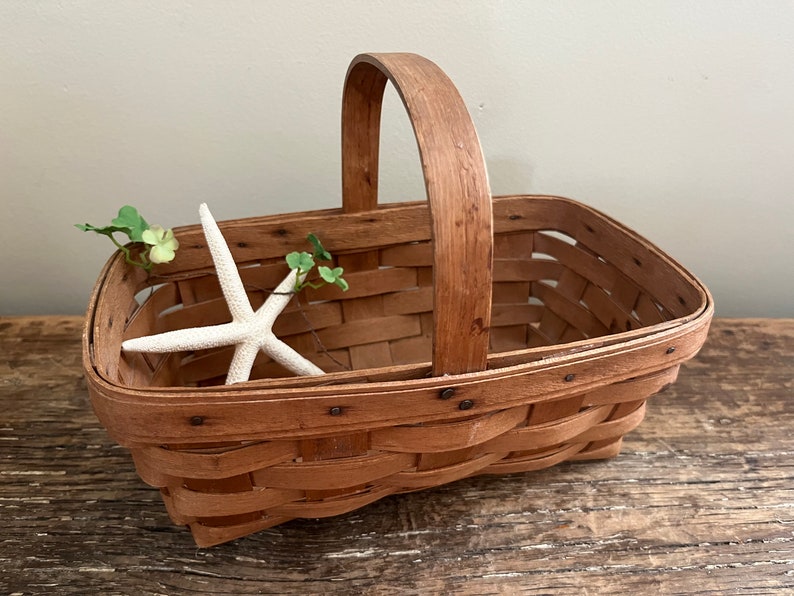 May include: A brown woven basket with a wooden handle. The basket is empty and has a white starfish and green leaves inside.