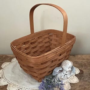 May include: A brown woven basket with a single handle. The basket is sitting on a white doily with a small porcelain angel figurine and purple flowers.