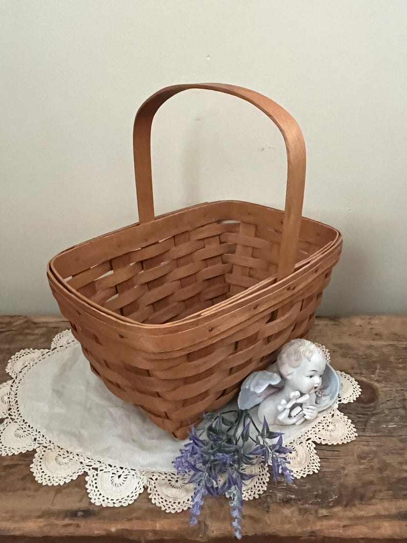 May include: A brown woven basket with a single handle. The basket is sitting on a white doily with a small porcelain angel figurine and purple flowers.