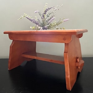 May include: A small wooden stool with a shelf. The stool is made of light brown wood and has a simple design. It is sitting on a black surface.