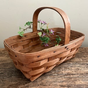 May include: A small, rectangular, wooden basket with a woven design and a handle. The basket is filled with a sprig of purple flowers.