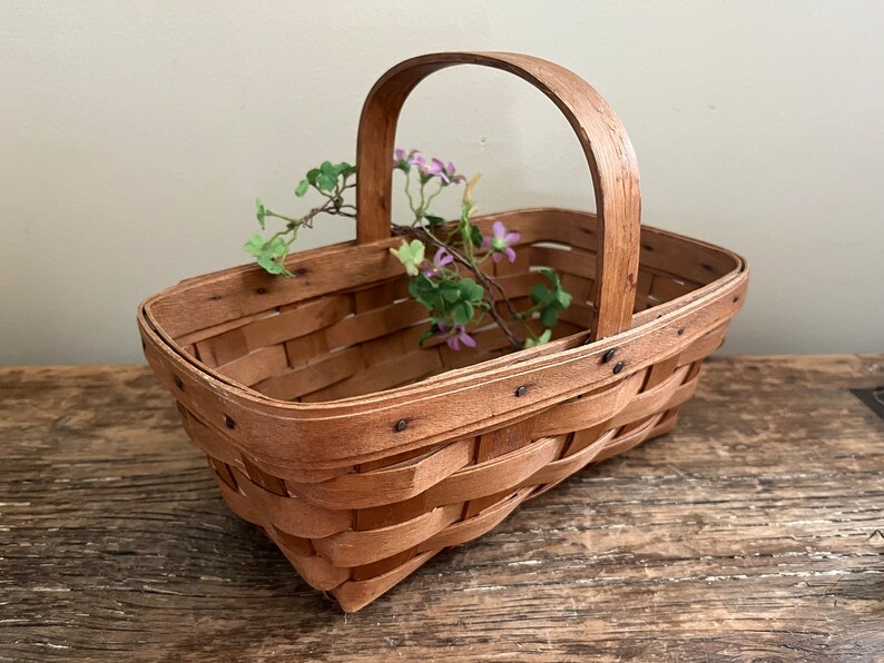 May include: A small, rectangular, wooden basket with a woven design and a handle. The basket is filled with a sprig of purple flowers.