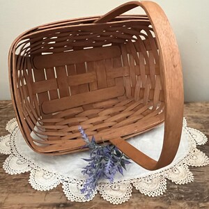 May include: A natural woven wood basket with a single handle. The basket is empty and sitting on a white doily.