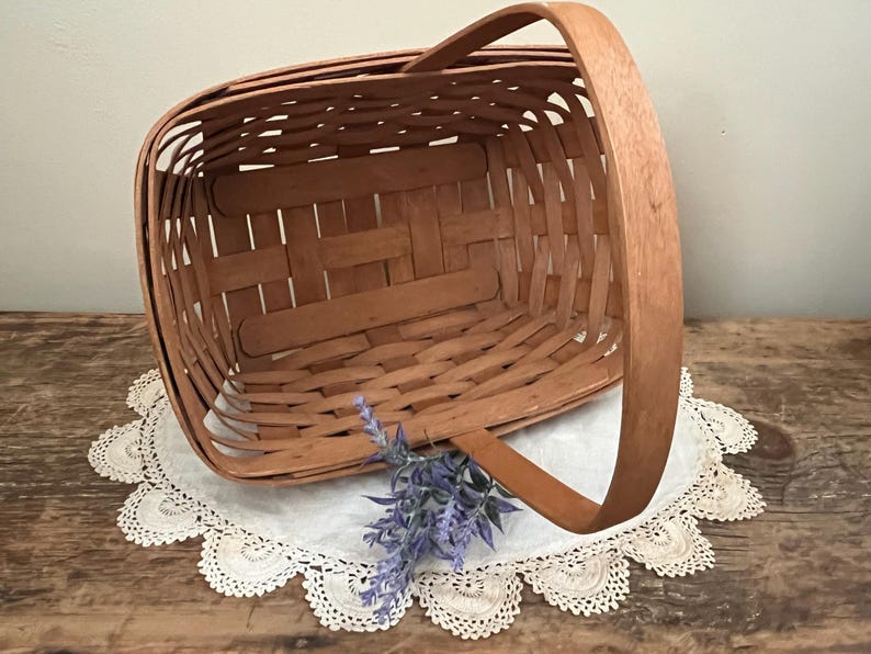 May include: A natural woven wood basket with a single handle. The basket is empty and sitting on a white doily.