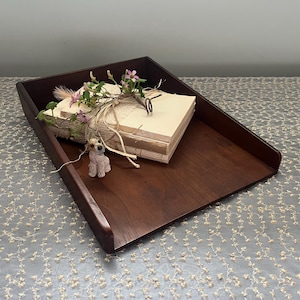 May include: A dark brown wooden tray with a stack of papers and a small stuffed animal on top. The tray is sitting on a white lace tablecloth.