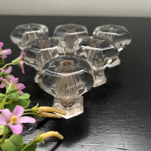 May include: Six clear glass cabinet knobs with a faceted design. The knobs have a rounded top and a hexagonal base. The knobs are arranged in a group, with a few sprigs of pink flowers in the foreground.