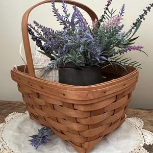 May include: A brown wicker basket with a handle, filled with a bouquet of purple and pink lavender flowers. The basket is sitting on a white crocheted doily.