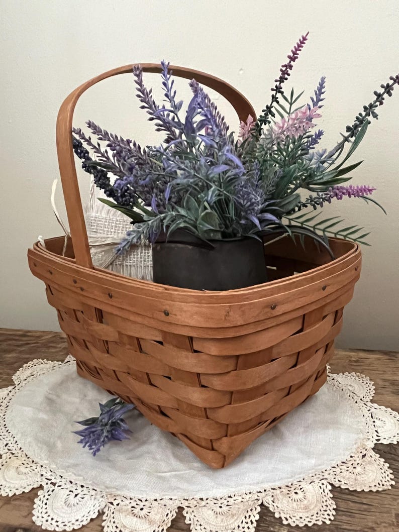 May include: A brown wicker basket with a handle, filled with a bouquet of purple and pink lavender flowers. The basket is sitting on a white crocheted doily.
