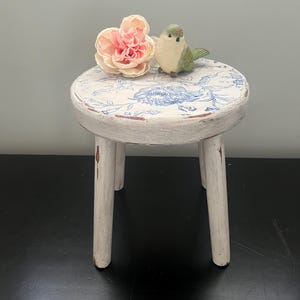 May include: A small, round, white wooden stool with a distressed finish. The top features a blue floral pattern. A pink flower and a small green and white bird figurine sit on top. The stool has three legs.