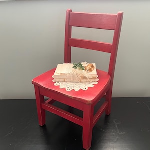 May include: A red wooden chair with a single back slat and a small stack of books on the seat. The chair is sitting on a black surface.
