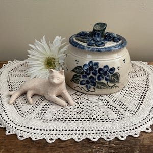 May include: A ceramic jar with a blue floral design and a matching lid sits on a white crocheted doily. A small, light pink cat figurine and a white daisy are also on the doily. The jar is a cream color with blue accents.