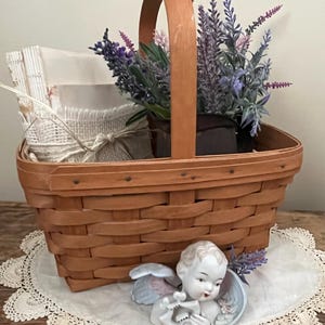 May include: A brown wicker basket with a handle filled with lavender flowers and a porcelain cherub figurine. The basket is sitting on a white crocheted doily.