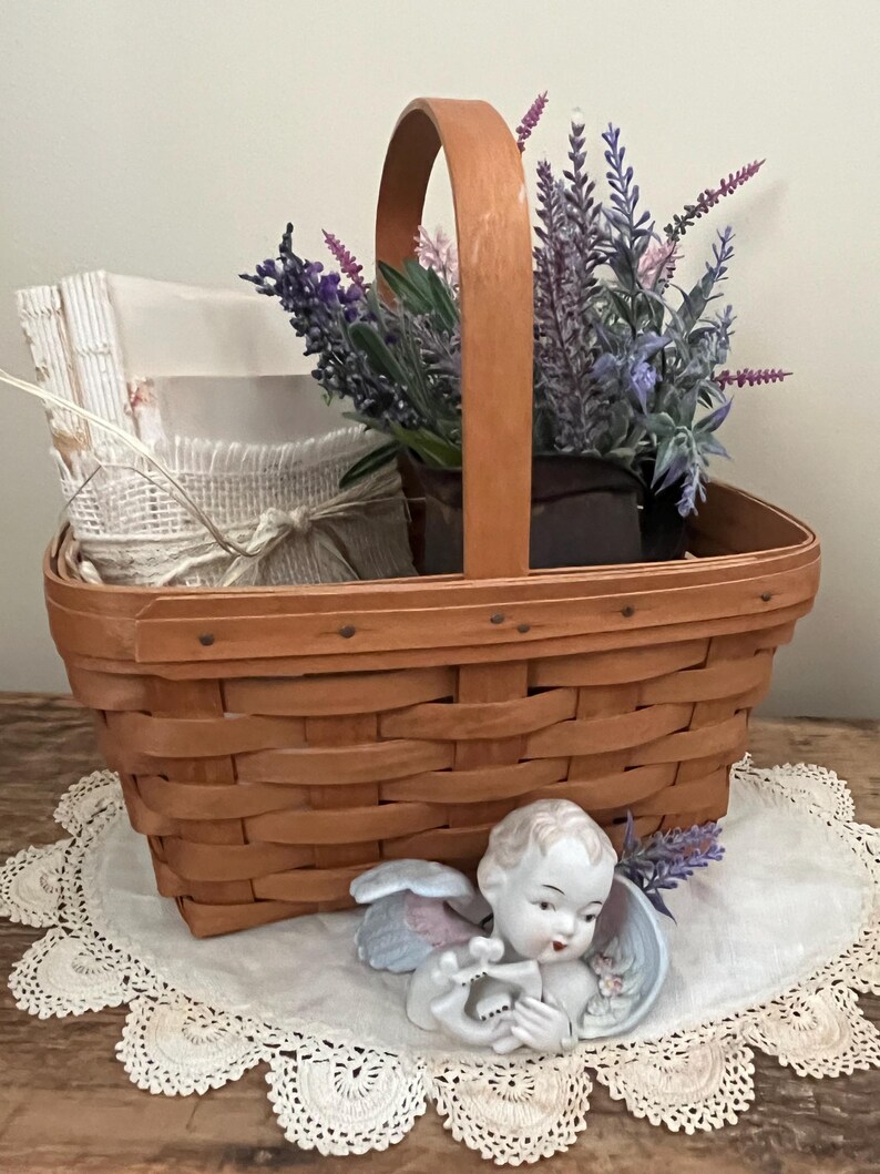 May include: A brown wicker basket with a handle filled with lavender flowers and a porcelain cherub figurine. The basket is sitting on a white crocheted doily.