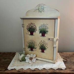 May include: A small, off-white wooden cabinet with a decorative floral design on the top. The cabinet door features illustrations of potted herbs. A small, green bird figurine and a white flower sit on a lace doily in front of the cabinet.
