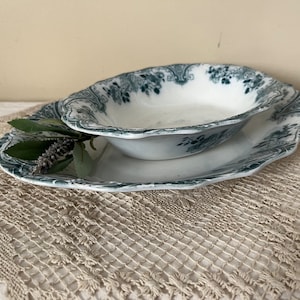May include: A vintage white porcelain serving dish with a green floral pattern. The dish is oval shaped and has a scalloped edge. It is sitting on a white crocheted doily.