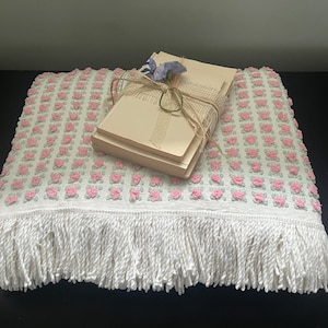 May include: A white crocheted throw blanket with a pink floral pattern and a white fringe border. A stack of wrapped paper is on top of the blanket.