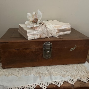 May include: A rectangular wooden box with a dark brown finish and a central metal latch. Resting on top is a stack of off-white paper pages tied with white string and topped with a white artificial flower. The box sits on a white lace doily.