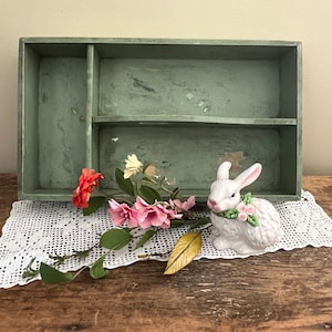 May include: A white ceramic rabbit figurine with pink flowers around its neck sits on a white crocheted doily. The rabbit is in front of a green wooden box with three compartments.