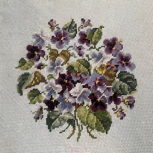 May include: Needlepoint of a bouquet of violets. The flowers are in shades of purple, lavender, and white, with green leaves. The needlepoint is on a light gray background.