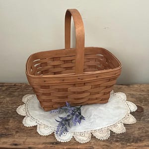 May include: A natural woven wood basket with a rectangular shape and a single handle. The basket is sitting on a white doily with a purple flower.