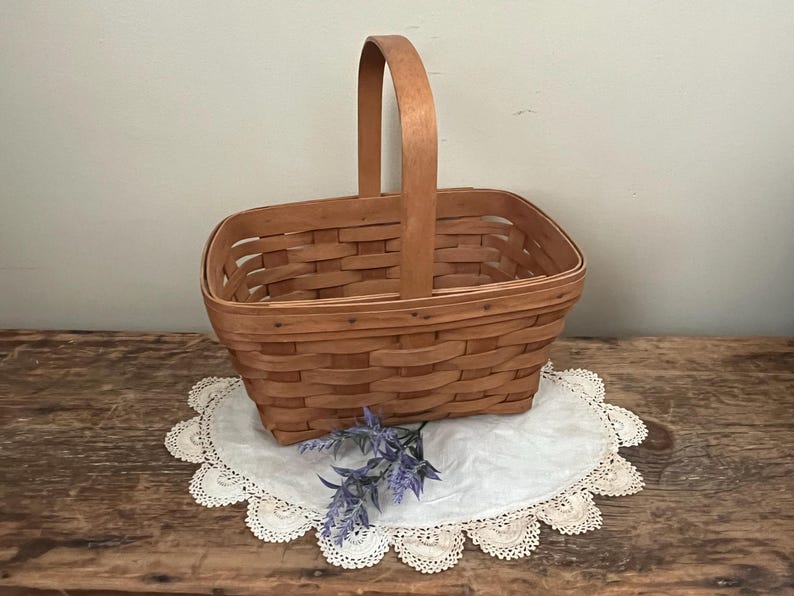 May include: A natural woven wood basket with a rectangular shape and a single handle. The basket is sitting on a white doily with a purple flower.